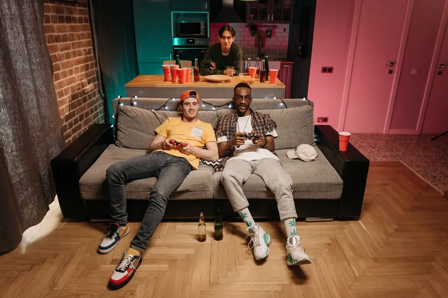 Two friends on a grey couch holding game controllers under string lights, with a third friend at the kitchen counter behind them and red cups on the bar
