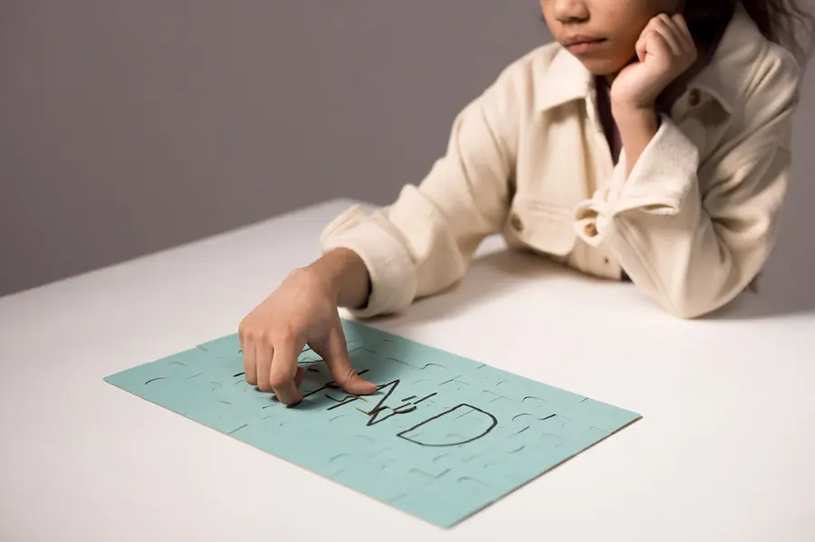 A child in a cream jacket rests their chin on one hand and traces letters drawn on a mint-green jigsaw puzzle