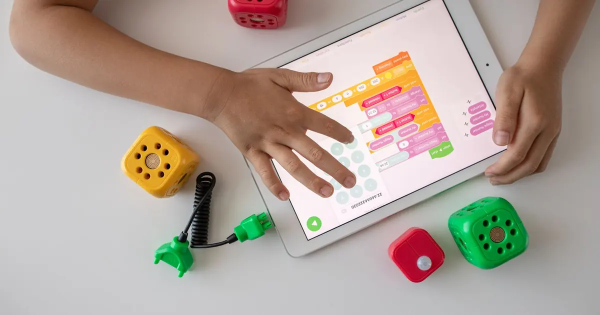 A child's hands on a white tablet showing colourful block-coding tiles, with toy dice-shaped robots on the table