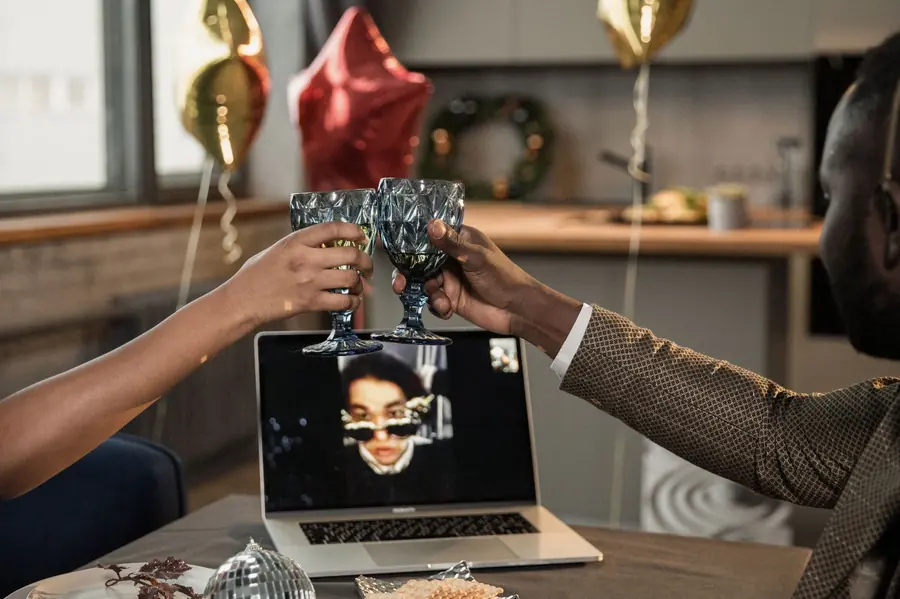 Two people clink patterned glasses in front of a laptop video call, with foil balloons and a wreath in the background