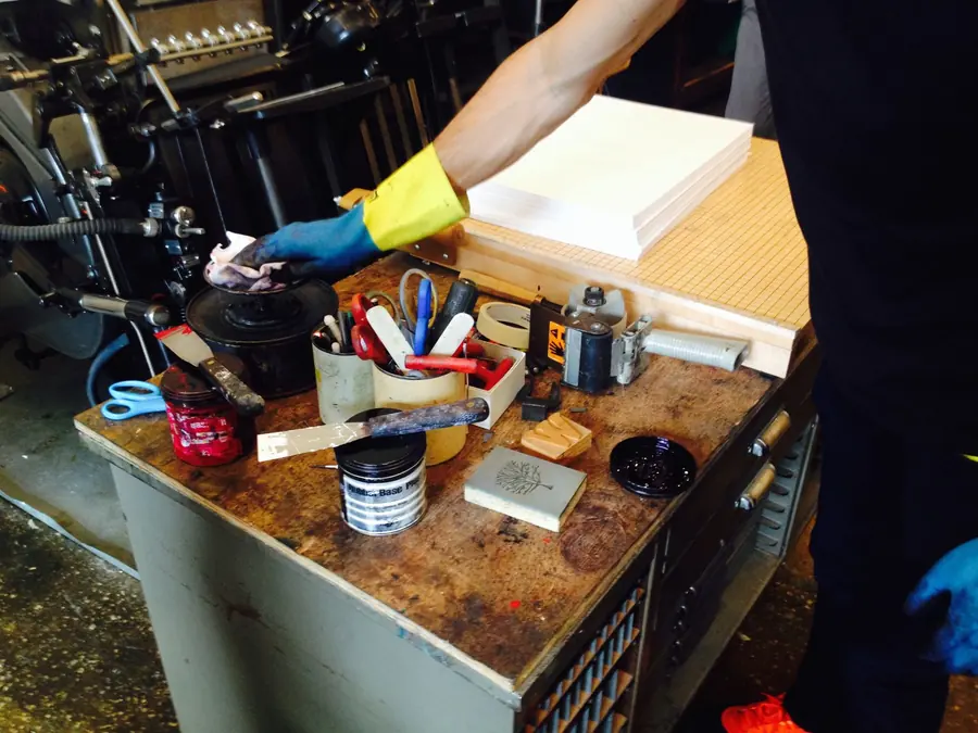 A print-shop workbench with a stack of white paper, blue-handled scissors, packing tape, brushes and tins of ink